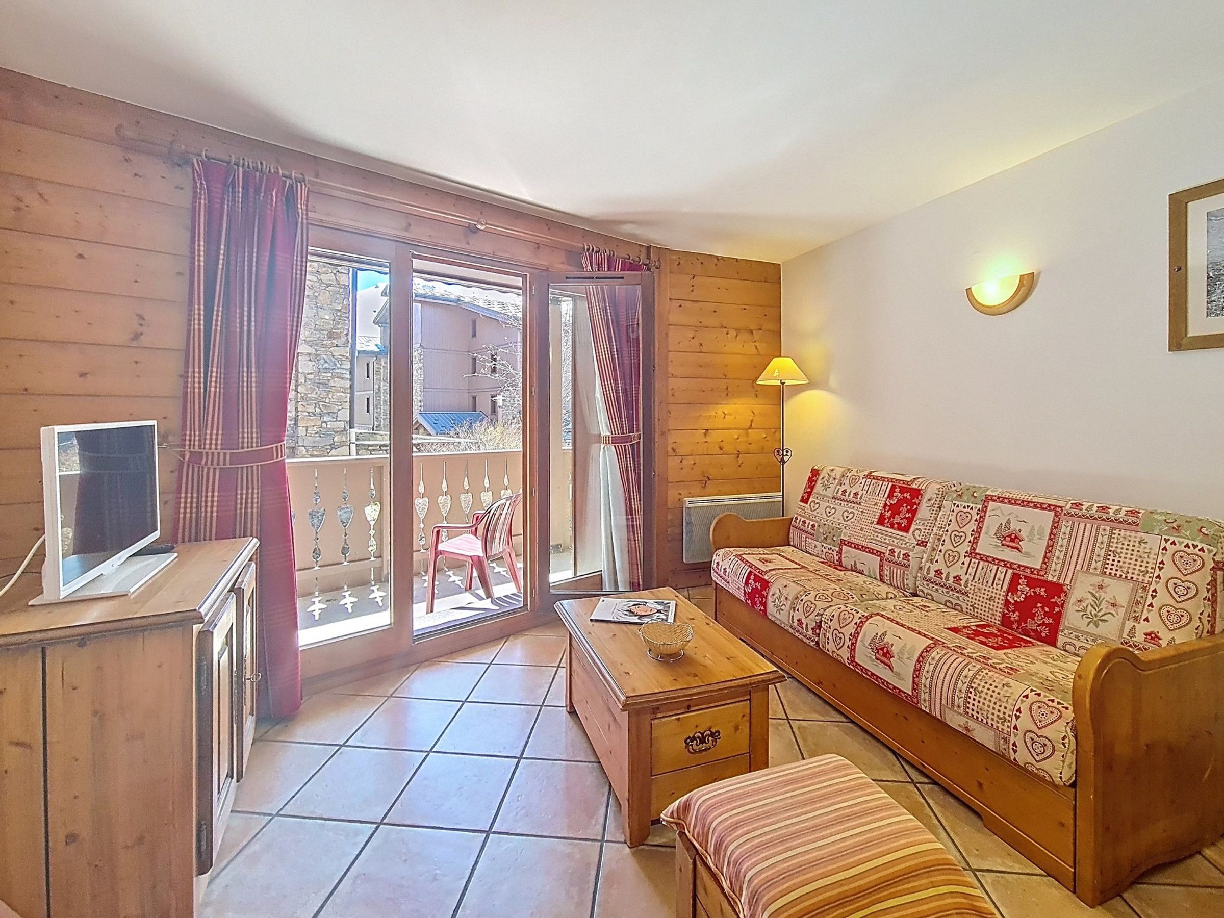 A cozy room with a wooden floor, a sofa with a patterned cover, a television on a wooden cabinet, and a view through a glass door to the outside.
