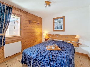 A room with a wooden wall, a bed with a blue patterned bedspread, a tray with cups and a kettle, a lamp, and a framed picture on the wall. - Apartment near slopes, balcony (Les Menuires)