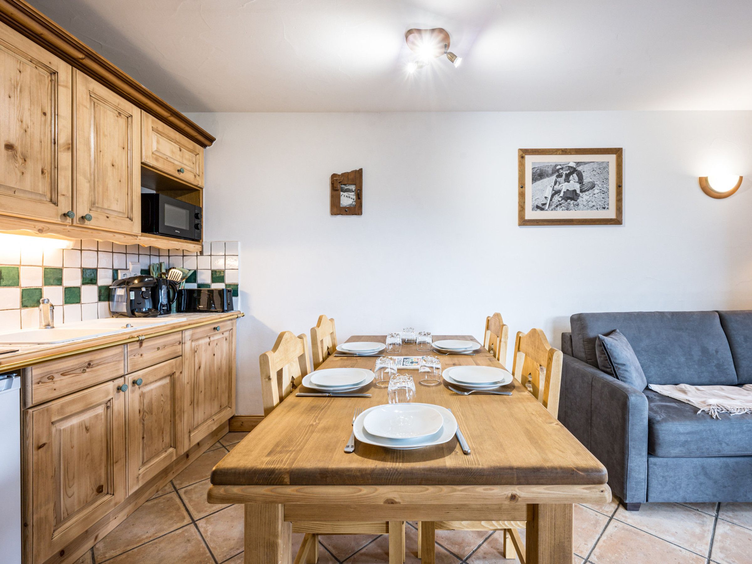 Un coin repas avec une table en bois dressée pour quatre personnes, une cuisine avec des meubles en bois et un canapé gris.