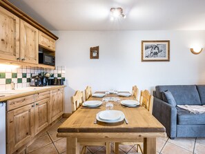 A dining area with a wooden table set for four, a kitchen with wooden cabinets, and a grey sofa. - Spacious apartment near slopes - 6 guests (Les Menuires)