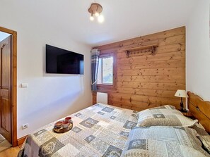 A bedroom with a wooden headboard, a flat-screen TV mounted on the wall, a window with curtains, and a quilted bedspread. - Spacious apartment, near slopes - 4 guests (Les Menuires)
