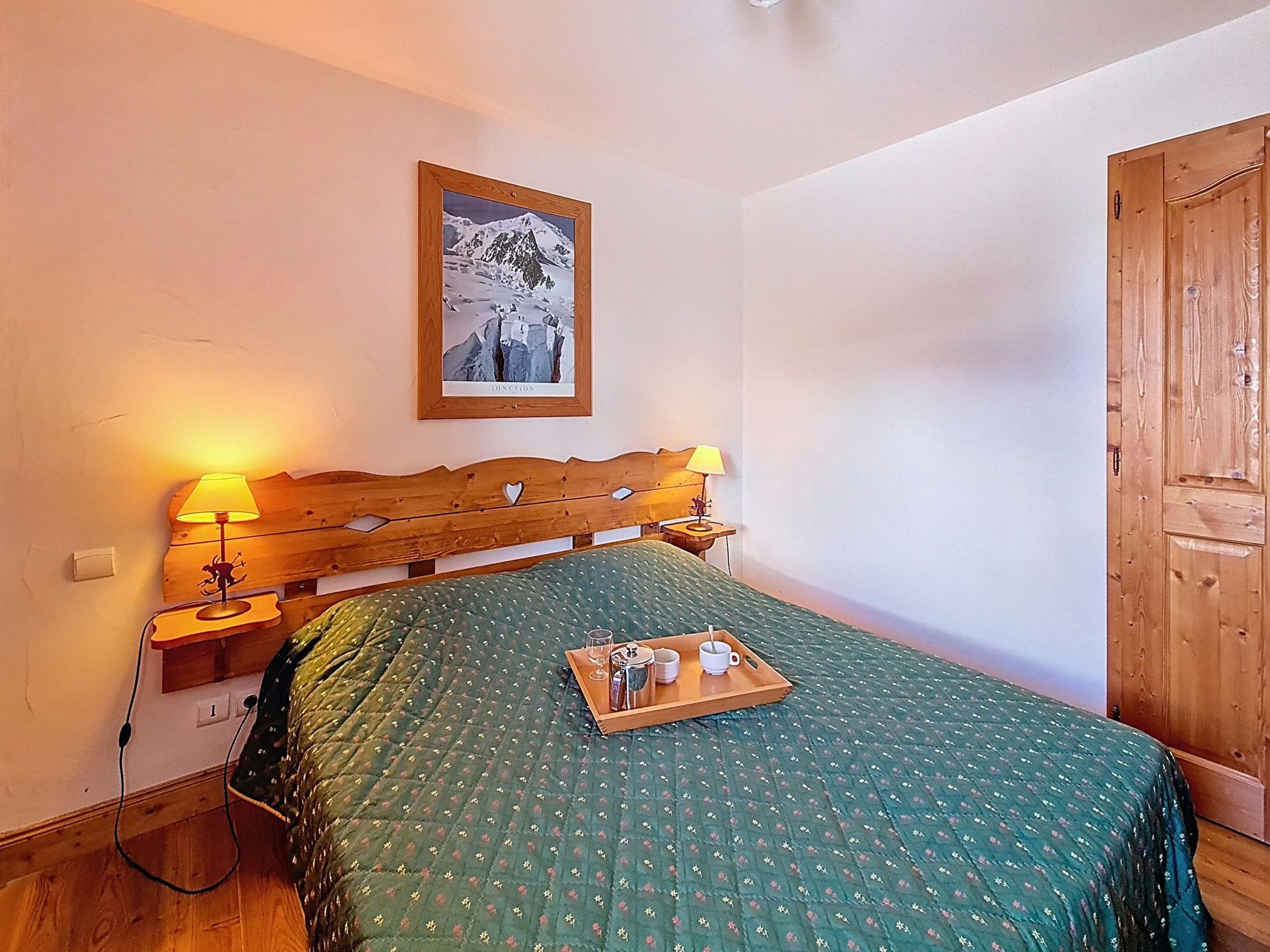 Quarto com cabeceira de madeira, colcha acolchoada verde, mesa de cabeceira com luminária e bandeja com xícaras e um copo.