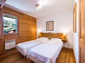 A room with wooden walls and a window overlooking a mountainous landscape. There are two beds with white bedding, a bedside table with a lamp, and a wall-mounted picture. - Spacious apartment with balcony - 4 guests (Les Menuires)