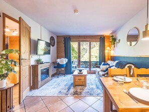 A cozy living room with a fireplace, a television mounted on the wall, a dining area with a table set for two, and a large window offering a view of the outdoors. - Spacious 2-room apartment with balcony, near slopes, parking included (Les Menuires)