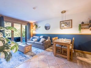 A living room with a sofa, coffee table, and dining table. There are windows, a plant, and a wall-mounted mirror. - Spacious 2-room apartment with balcony, near slopes, parking included (Les Menuires)