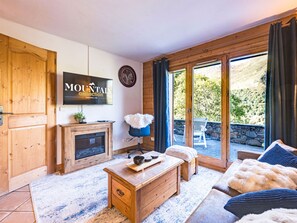 A cozy living room with a fireplace, a television, and a view of the outdoors. - Spacious 2-room apartment with balcony, near slopes, parking included (Les Menuires)