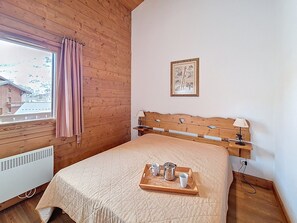 Ein Schlafzimmer mit hölzernem Kopfteil, einem Fenster mit Vorhängen, einem Nachttisch mit Lampe sowie einer Ablage mit Tassen und einer Kaffeekanne.