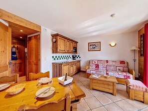 A cozy living area with a wooden dining table set for two, a sofa with a patterned cover, and a kitchen with wooden cabinets and a checkered backsplash. - Spacious and bright apartment near slopes (Les Menuires)