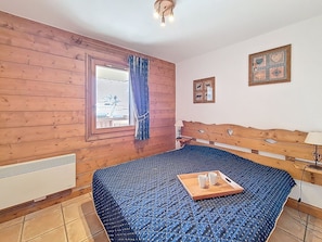 Ein Zimmer mit einem Bett, das ein Holz-Kopfende hat, einem Vorhangfenster, einem Heizkörper und einem kleinen Tisch mit Tassen.