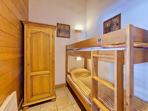 Un lit superposé avec une tête de lit en bois et un lit simple avec un oreiller à rayures. Une armoire en bois est adossée au mur.