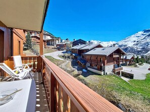 A balcony with a table and chairs, overlooking a mountain village with wooden buildings and a clear blue sky. - Spacious apartment - 8 guests. Near the slopes (Les Menuires)
