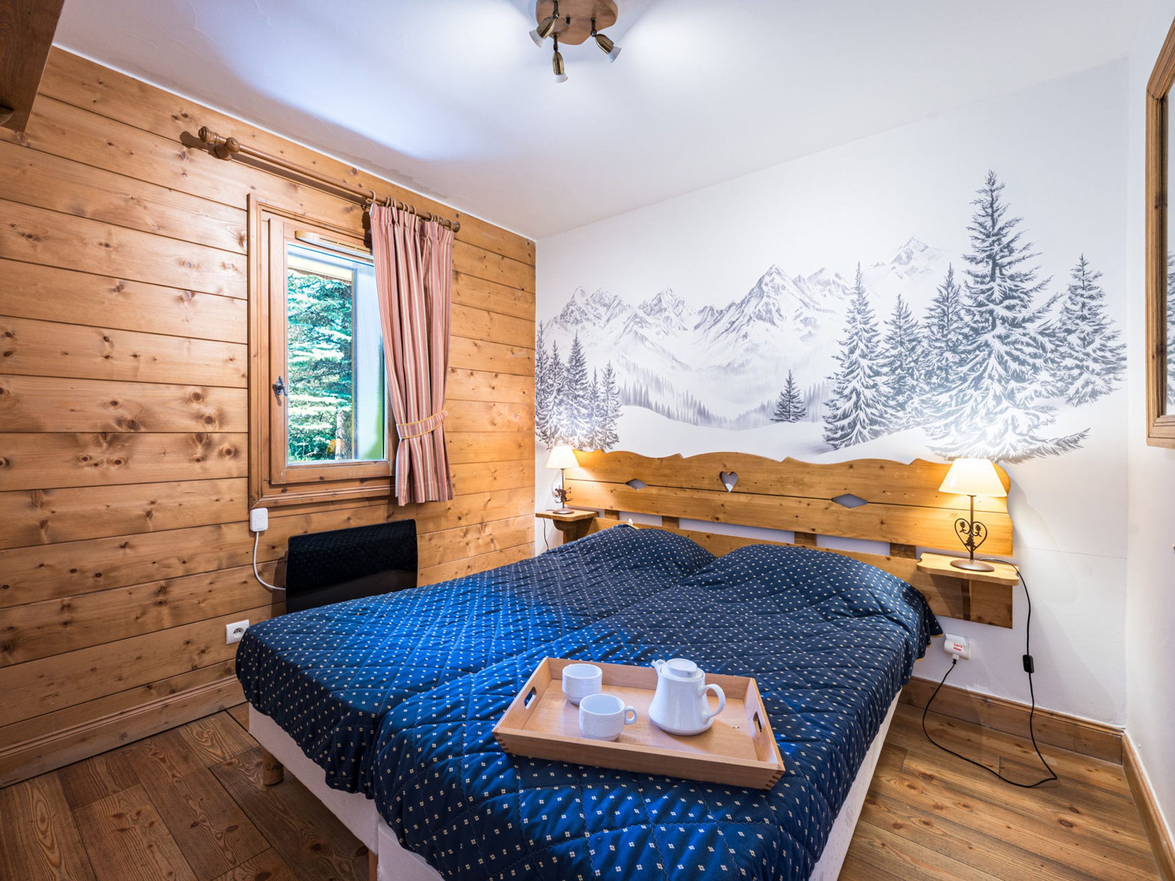 Une chambre à coucher avec une tête de lit en bois, un lit recouvert d’une courtepointe bleue, une fenêtre avec des rideaux, une fresque murale représentant une montagne enneigée et une table de chevet avec une lampe.