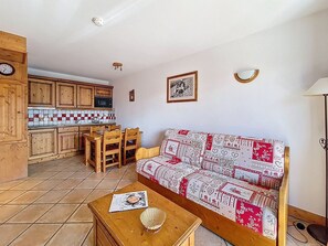 A cozy living room with a patterned sofa, a wooden table with a basket, and a tiled kitchen area with wooden cabinets. - Spacious apartment near slopes - 4 guests (Les Menuires)