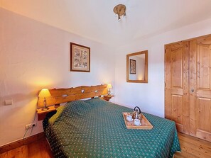 Un dormitorio con cabecera de madera, una cama con colcha verde estampada, una mesita de noche con lámpara, una bandeja con tetera y tazas, y una puerta de madera.