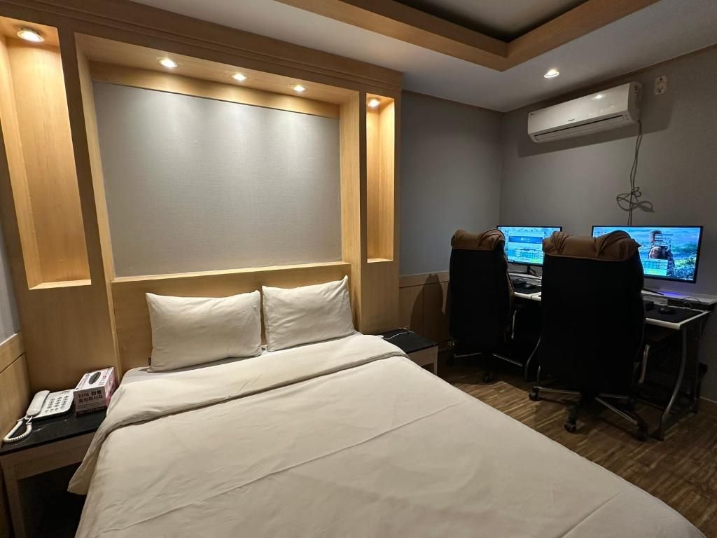 A hotel room with a bed, a desk with two monitors, a telephone, and an air conditioning unit.