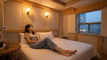 Una habitación de hotel con cama, mesita de noche, lámpara y ventana con vistas a edificios.