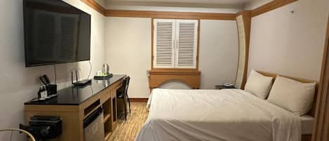 A bedroom with a bed, a desk, a television, and a window with shutters.