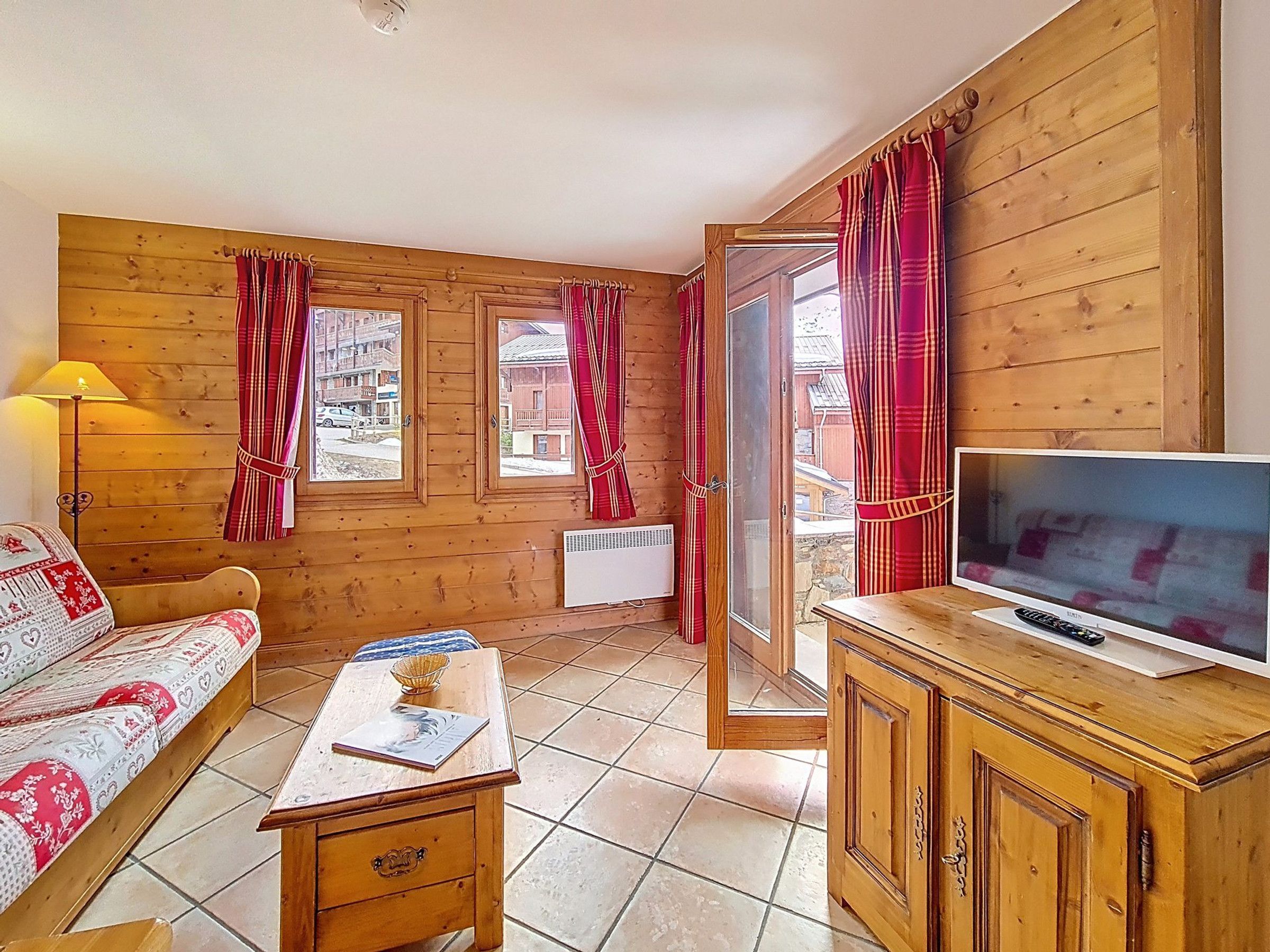 Una habitación acogedora con detalles de madera, una cama con ropa de cama estampada, una mesita con un cuenco, un televisor sobre un mueble de madera y una ventana con cortinas.
