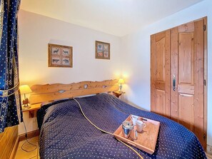 Une chambre à coucher avec une tête de lit en bois, deux tableaux accrochés au mur, une porte en bois et une table de chevet sur laquelle se trouvent une lampe et un plateau avec une bouilloire et des tasses.