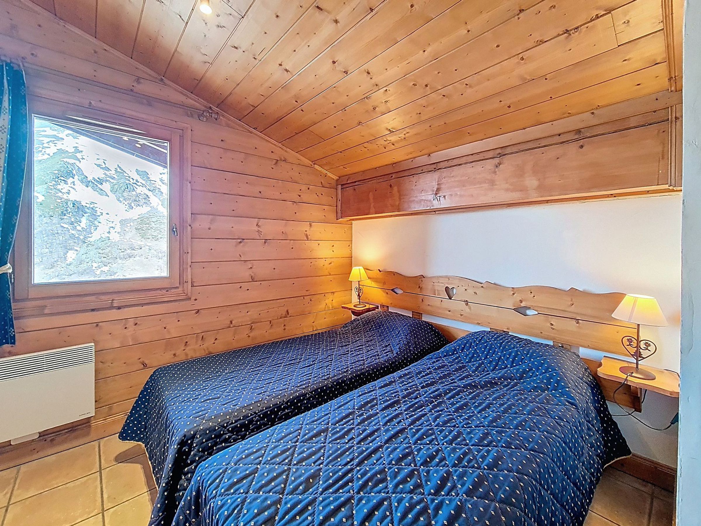 A room with wooden walls and ceiling, two beds with blue bedding, a window with a view, and a heater on the wall.