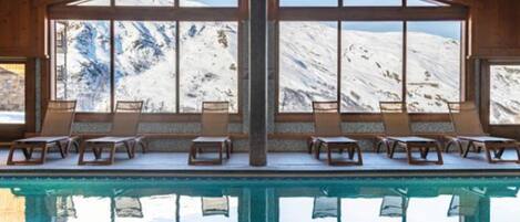 Zona de piscina con sillones de descanso y ventanas amplias con vistas a montañas nevadas.