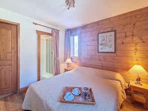 Une pièce avec un lit, un mur en bois, une fenêtre avec des rideaux, un tableau encadré et un plateau avec des tasses et une théière posé sur le lit.