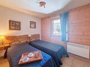 Ein Zimmer mit zwei Betten, einem fenster mit Vorhängen, einem Heizkörper und einem Tablett mit Tassen und einem Topf auf dem Boden.