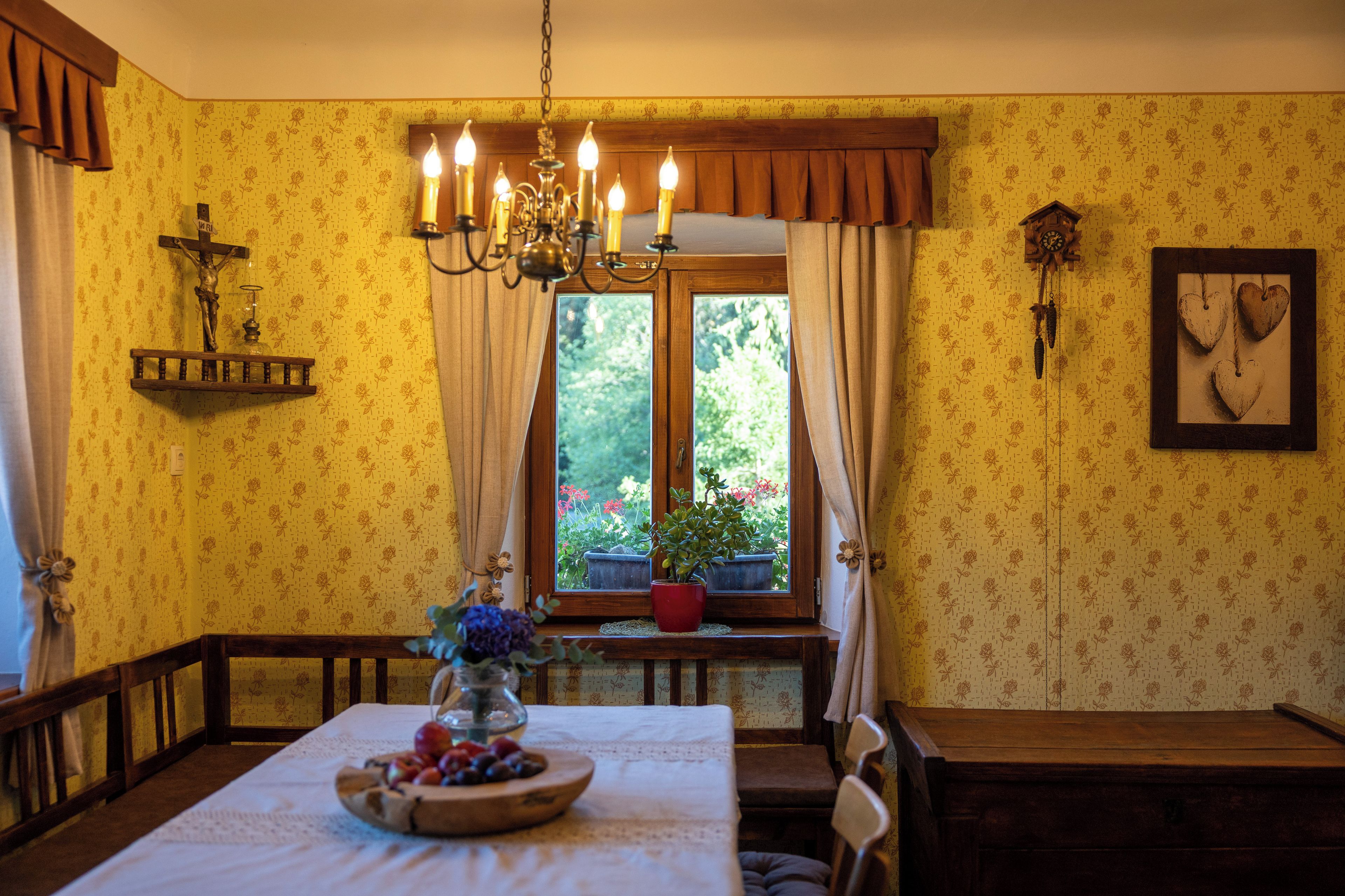 Un comedor con una mesa de madera, sillas, una araña, un crucifijo y una ventana que ofrece vistas a la vegetación.