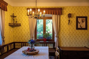 A dining room with a wooden table, chairs, a chandelier, a crucifix, and a window with a view of greenery.