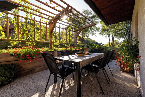 An outdoor patio with a wooden pergola, a table with chairs, and a variety of plants and flowers.