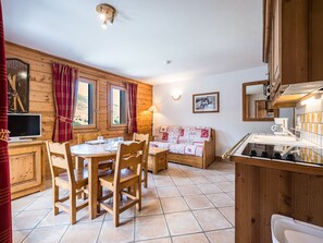 A wooden interior with a dining table, chairs, a sofa, a TV, and a kitchen area. - Cozy apartment - 4 guests (Les Menuires)