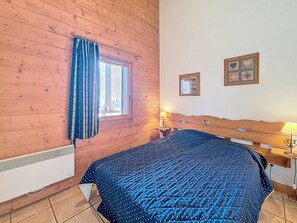 Ein Zimmer mit einer Wand aus Holzlatten, einem Fenster mit Vorhängen, einem Bett mit blauer Steppdecke, einem Nachttisch mit Lampe und zwei gerahmten Bildern an der Wand.