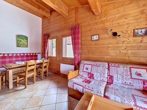 A wooden cabin interior with a dining table, chairs, a sofa with patterned cushions, and a wall-mounted light fixture. - Spacious apartment near slopes - 6 guests (Les Menuires)