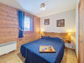Une chambre à coucher avec une tête de lit en bois, une fenêtre avec des rideaux, un lit recouvert d’une couette bleue, une table de chevet avec une lampe et un plateau avec des tasses et une théière.