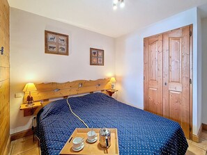 Ein Schlafzimmer mit einem hölzernen Kopfteil, einem Bett mit blauer Steudecke, einem Nachttisch aus Holz mit einer Lampe, einer Holztür und zwei gerahmten Bildern an der Wand.