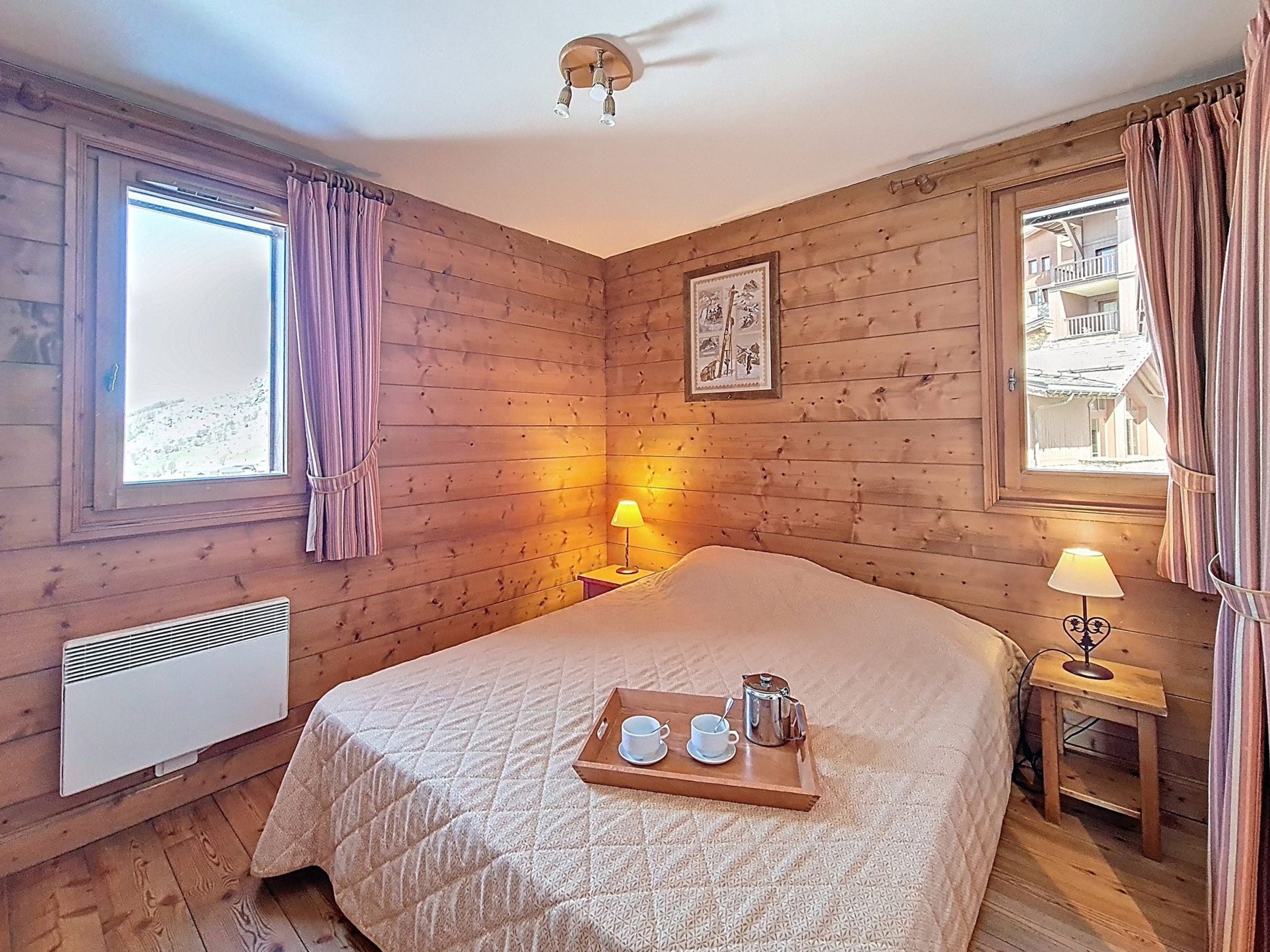 Ein Zimmer mit Holzwänden, einem Bett, zwei Nachttischlampen, einem Fenster mit Vorhängen, einem Heizkörper und einem Tablett mit Tassen und einer Teekanne.