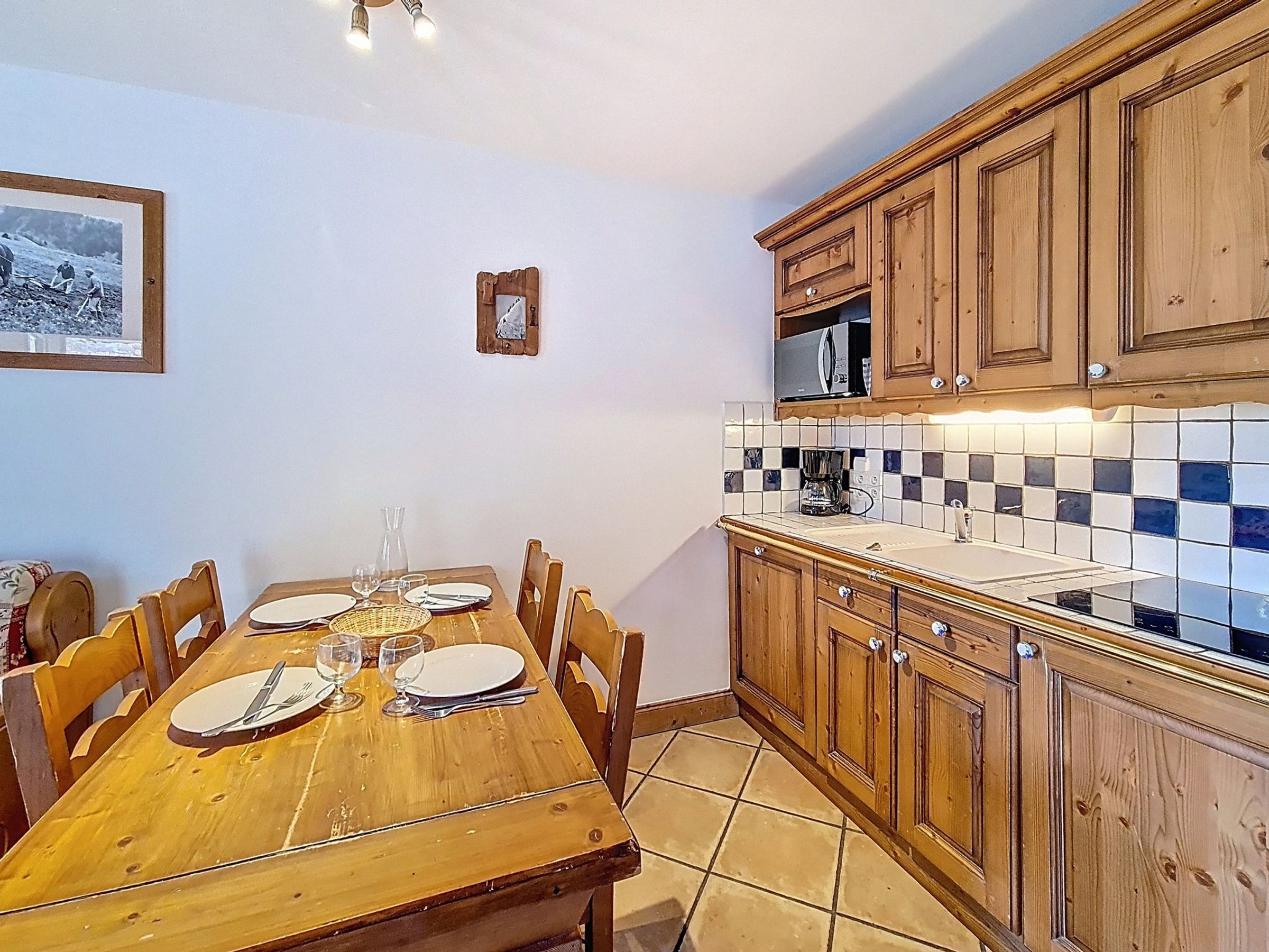 A dining area with a wooden table set for four, a kitchen with wooden cabinets, a microwave, and a tiled backsplash.