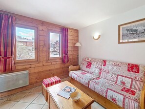 A cozy room with a wooden interior, a patterned sofa, a small table with a book, and a window with curtains. - Spacious apartment near slopes - 6 guests (Les Menuires)