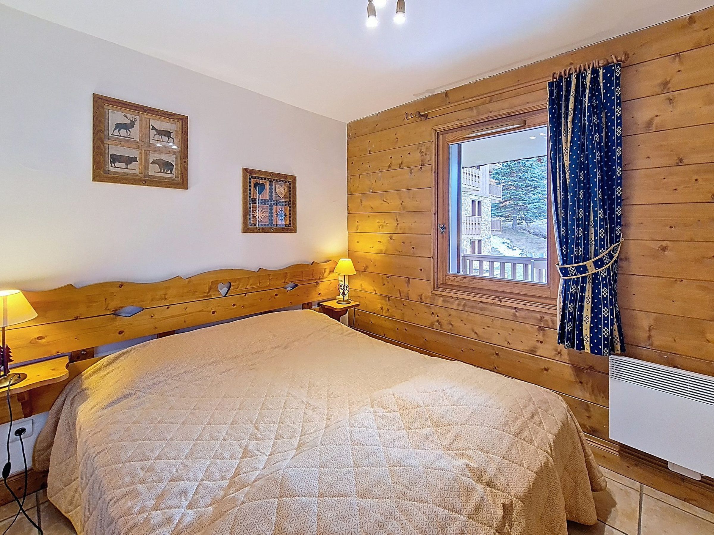 A bedroom with a wooden headboard, a bed with a quilt, a bedside lamp, a window with curtains, and two framed pictures on the wall.