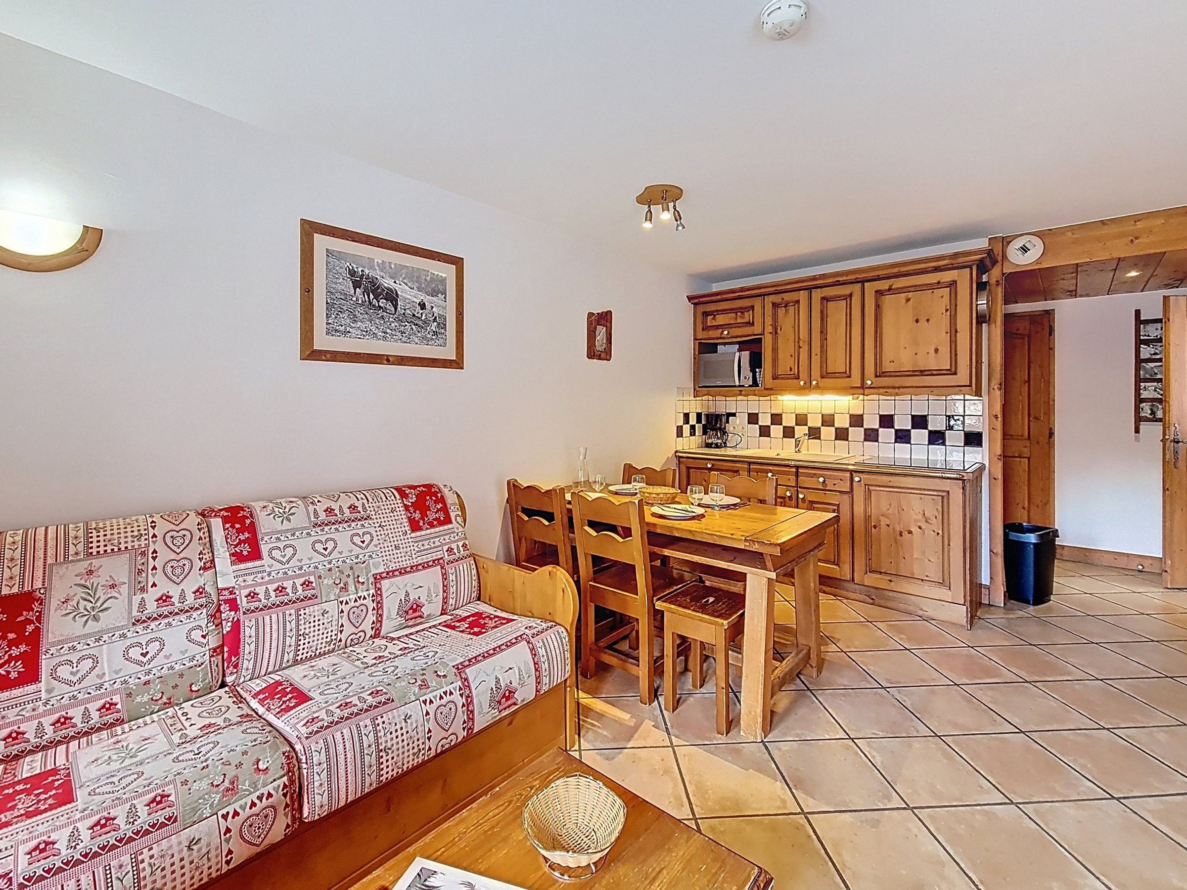 A cozy living room with a patterned sofa, a wooden dining table with chairs, and a kitchen area with wooden cabinets and a tiled backsplash.