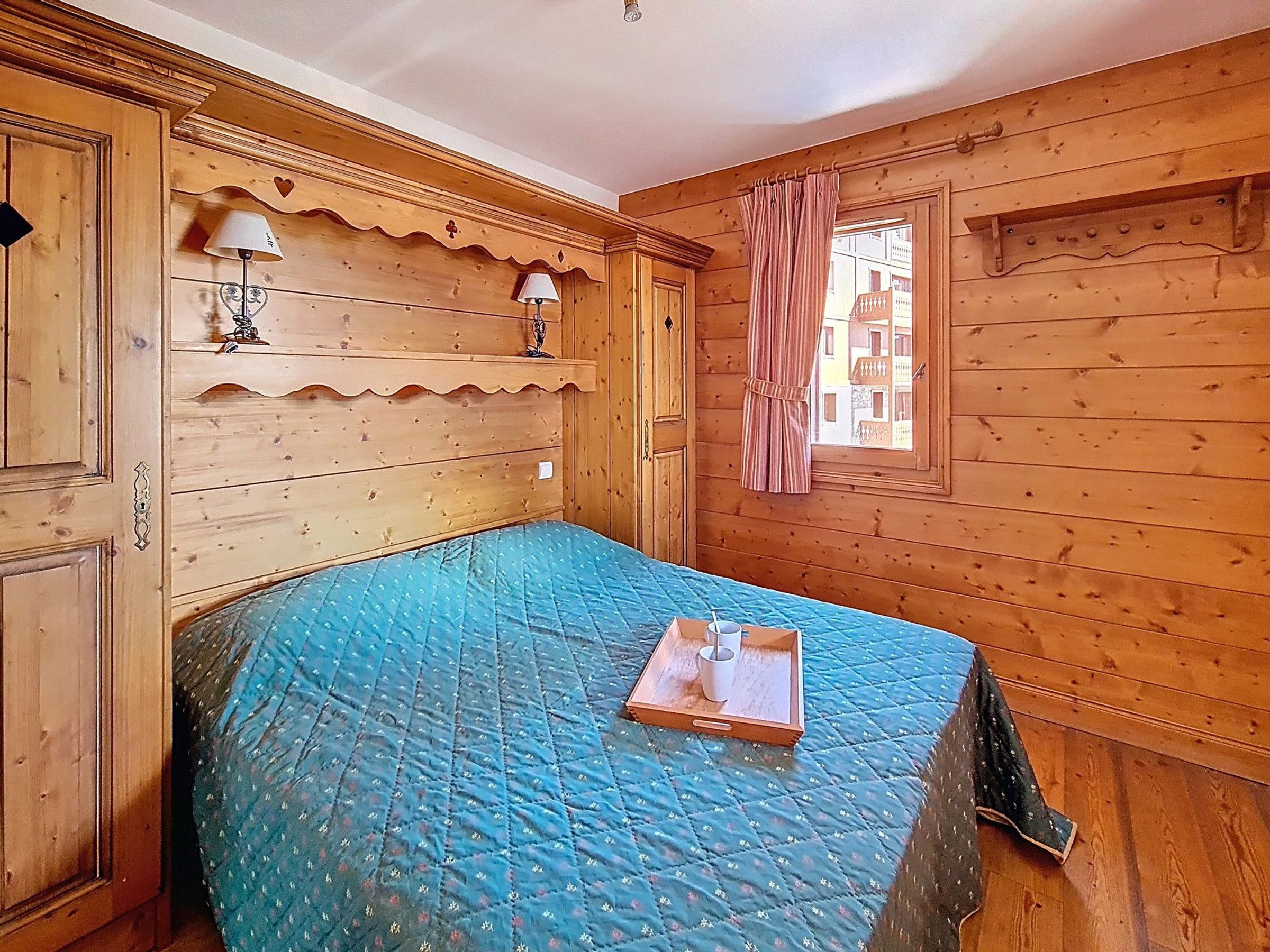 Ein Zimmer mit Holzboden, einem Bett, zwei Wandlampen, einem Fenster mit Vorhängen und Blick auf Gebäude.