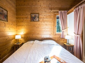 A wooden cabin room with a bed, two bedside tables, a window with curtains, and a painting on the wall. - Spacious apartment near slopes - 6 guests (Les Menuires)