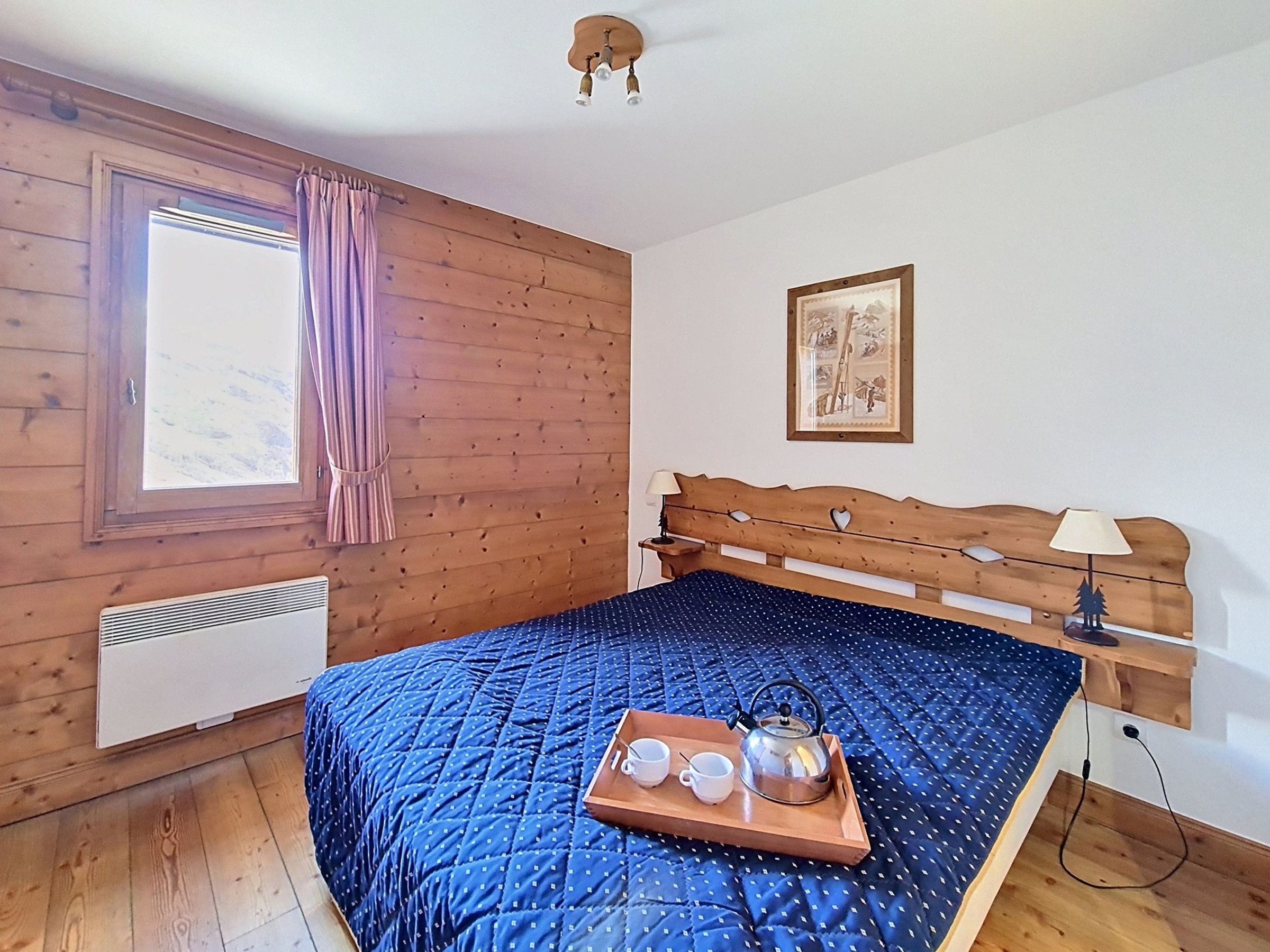 Ein Zimmer mit einem hölzernen Bett, einem Vorhangfenster, einem Wandbild und einem Nachttisch mit Lampe.