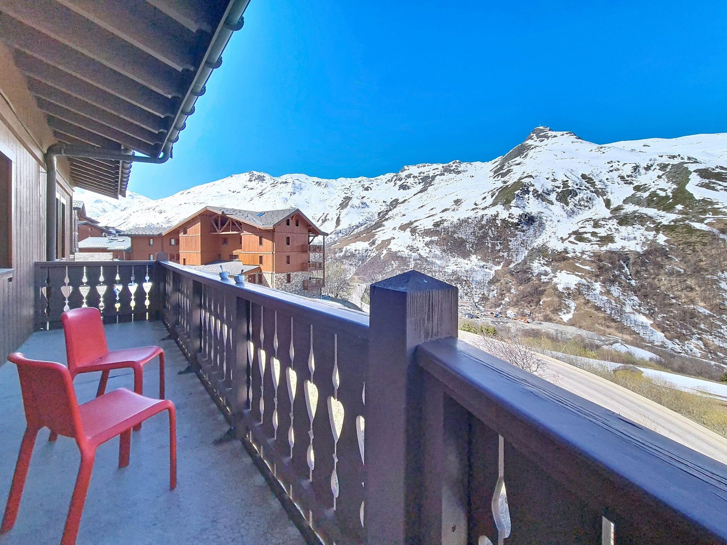 Een balkon met een houten leuning, twee rode stoelen en uitzicht op besneeuwde bergen.
