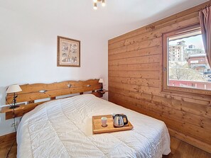 A hotel room with a wooden headboard, a bed with a white quilt, a bedside table with a lamp, a tray with a cup and a kettle, and a window with a view of buildings. - Apartment near slopes, balcony - 6 guests (Les Menuires)