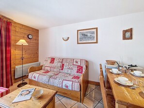 A cozy room with a wooden sofa, a dining table set with plates and glasses, a lamp, and a wall clock. - Apartment near slopes, balcony - 6 guests (Les Menuires)