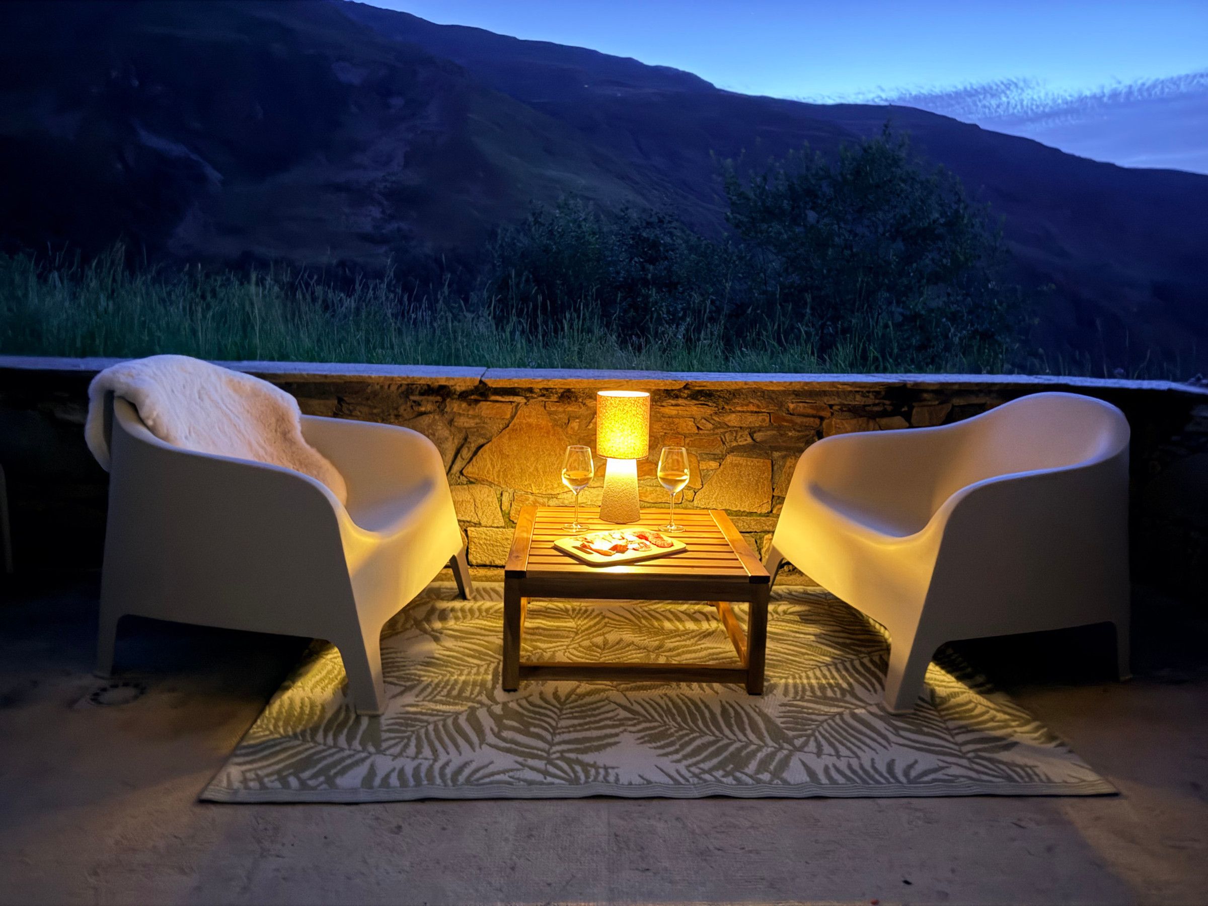 Un área de estar al aire libre con dos sillas, una mesa, un muro de piedra y vistas a las colinas.