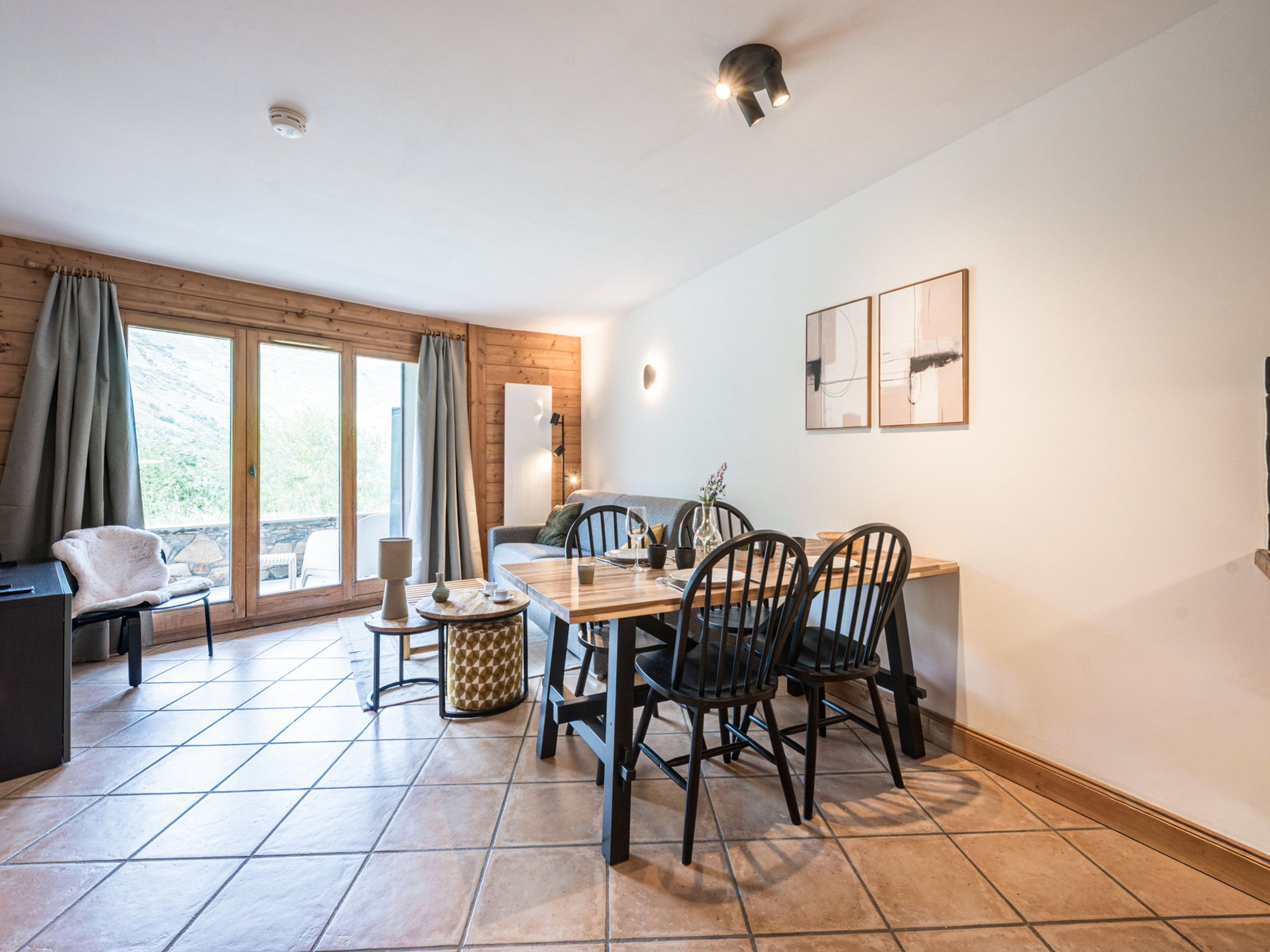 A dining area with a wooden table, four chairs, and a sofa. There are curtains on the windows, and a lamp on the wall.