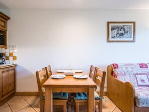 A dining area with a wooden table set for two, a kitchen area with a coffee machine, and a patterned sofa. - Bright apartment near slopes - 4 guests (Les Menuires)
