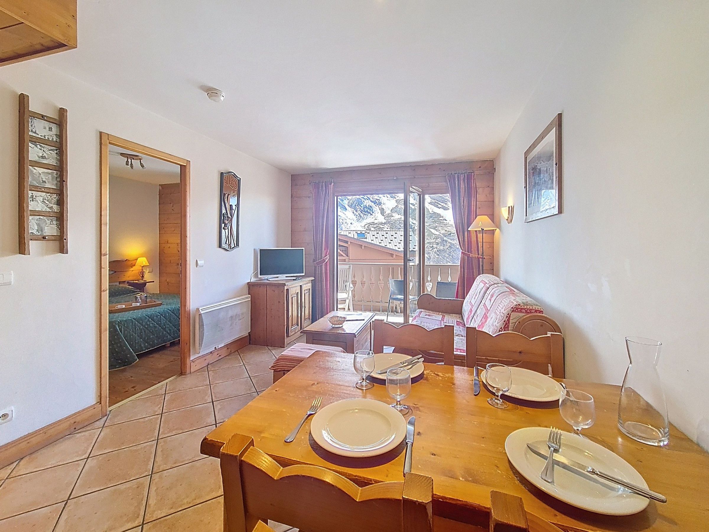 A dining area with a wooden table set for four, a television on a cabinet, and a balcony with a view of mountains.